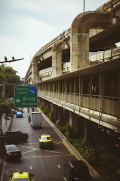 A busy urban road is flanked by an elevated concrete structure, possibly a metro or railway track. Several vehicles, including cars, taxis, and motorbikes, navigate the street. A prominent green road sign provides directions to a hospital and police station in both Thai and English. The scene is surrounded by greenery, and the atmosphere feels overcast.