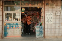 A small local shop with a variety of colorful items displayed, including toys, water bottles, and other goods. A person wearing a mask and an orange jacket stands at the entrance, surrounded by various wares. There are signs with Chinese characters on the door and exterior walls.