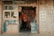 A small local shop with a variety of colorful items displayed, including toys, water bottles, and other goods. A person wearing a mask and an orange jacket stands at the entrance, surrounded by various wares. There are signs with Chinese characters on the door and exterior walls.
