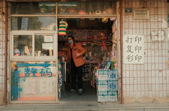 A small local shop with a variety of colorful items displayed, including toys, water bottles, and other goods. A person wearing a mask and an orange jacket stands at the entrance, surrounded by various wares. There are signs with Chinese characters on the door and exterior walls.
