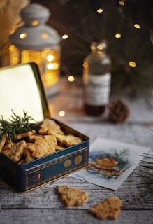 A festive setting features a tin box filled with tree-shaped cookies placed on a wooden surface. In the background, a softly lit lantern adds warmth, complemented by Christmas lights. A glass bottle and a holiday card with an illustration accompany the arrangement, with pine branches and cones enhancing the seasonal atmosphere.