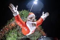 A large Santa Claus figure with arms raised stands amidst festive decorations at night. The figure is bathed in bright light from a streetlamp above, with greenery and strings of Christmas lights in the background. Raindrops are visible, adding a dynamic element to the scene.