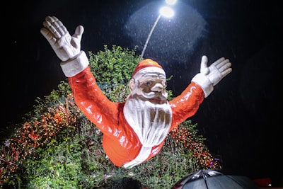 A large Santa Claus figure with arms raised stands amidst festive decorations at night. The figure is bathed in bright light from a streetlamp above, with greenery and strings of Christmas lights in the background. Raindrops are visible, adding a dynamic element to the scene.