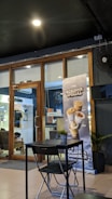 A cozy café interior featuring a modern, minimalist design with a small table and chair set. A prominent advertisement banner for 'Monsoon Special' beverages stands near a glass door, which gives a glimpse into another room with clothing on display. Potted plants add a touch of greenery.