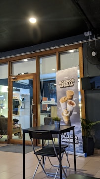 A cozy café interior featuring a modern, minimalist design with a small table and chair set. A prominent advertisement banner for 'Monsoon Special' beverages stands near a glass door, which gives a glimpse into another room with clothing on display. Potted plants add a touch of greenery.