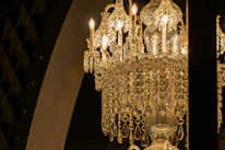 A luxurious crystal chandelier adorned with intricate designs and hanging crystals, illuminated by bright candle-shaped bulbs. The chandelier's golden glow casts a warm and elegant light, contrasting against a dark background with arch elements.