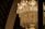 A luxurious crystal chandelier adorned with intricate designs and hanging crystals, illuminated by bright candle-shaped bulbs. The chandelier's golden glow casts a warm and elegant light, contrasting against a dark background with arch elements.