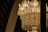 A luxurious crystal chandelier adorned with intricate designs and hanging crystals, illuminated by bright candle-shaped bulbs. The chandelier's golden glow casts a warm and elegant light, contrasting against a dark background with arch elements.