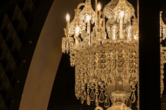 A luxurious crystal chandelier adorned with intricate designs and hanging crystals, illuminated by bright candle-shaped bulbs. The chandelier's golden glow casts a warm and elegant light, contrasting against a dark background with arch elements.