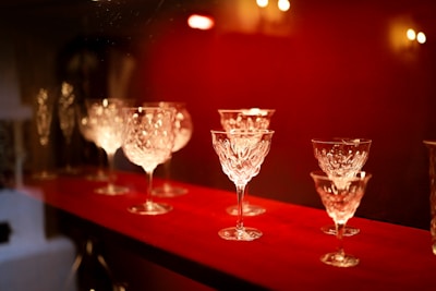 Elegant crystal wine glasses are displayed on a vibrant red shelf, with soft lighting creating a warm and inviting atmosphere. The intricate patterns on the glasses reflect the gentle light.