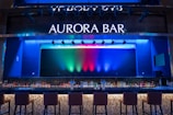 A stylish bar with a modern design featuring a large illuminated sign that reads 'Aurora Bar'. The bar has a sleek countertop with a variety of bottles neatly arranged behind it. Above, multicolored lights cast vibrant hues of blue, green, and red across the backdrop, creating a dynamic and lively atmosphere. High bar chairs line the front, inviting patrons to sit.