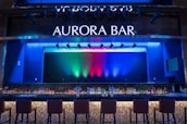 A stylish bar with a modern design featuring a large illuminated sign that reads 'Aurora Bar'. The bar has a sleek countertop with a variety of bottles neatly arranged behind it. Above, multicolored lights cast vibrant hues of blue, green, and red across the backdrop, creating a dynamic and lively atmosphere. High bar chairs line the front, inviting patrons to sit.