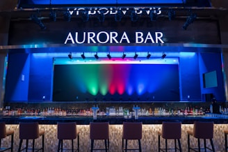 A stylish bar with a modern design featuring a large illuminated sign that reads 'Aurora Bar'. The bar has a sleek countertop with a variety of bottles neatly arranged behind it. Above, multicolored lights cast vibrant hues of blue, green, and red across the backdrop, creating a dynamic and lively atmosphere. High bar chairs line the front, inviting patrons to sit.