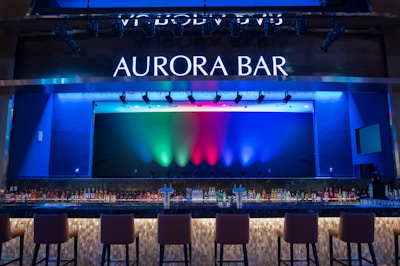 A stylish bar with a modern design featuring a large illuminated sign that reads 'Aurora Bar'. The bar has a sleek countertop with a variety of bottles neatly arranged behind it. Above, multicolored lights cast vibrant hues of blue, green, and red across the backdrop, creating a dynamic and lively atmosphere. High bar chairs line the front, inviting patrons to sit.