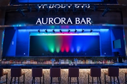 A stylish bar with a modern design featuring a large illuminated sign that reads 'Aurora Bar'. The bar has a sleek countertop with a variety of bottles neatly arranged behind it. Above, multicolored lights cast vibrant hues of blue, green, and red across the backdrop, creating a dynamic and lively atmosphere. High bar chairs line the front, inviting patrons to sit.