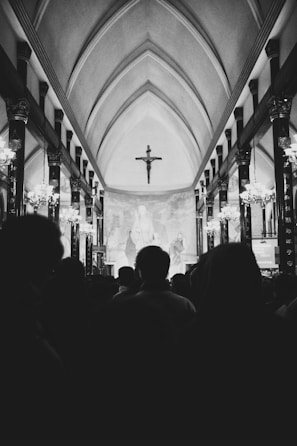 A large group of people is gathered inside a church with arched ceilings. The interior is decorated with multiple ornate columns and illuminated by hanging chandeliers. At the front, a crucifix is prominently displayed.