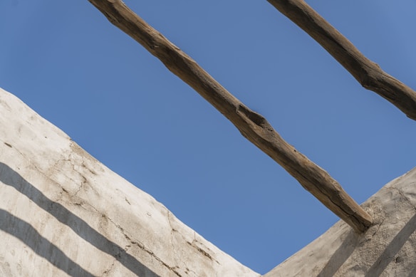 Horizontal wooden beams are laid across a section of a white, textured wall against a clear blue sky. Shadows of the beams create patterns on the wall.