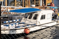 A white boat with a blue canopy is docked by a waterfront. The boat has several blue and white striped seats and a small cabin with windows. In the background, there is a building with wooden elements and stacked chairs. An orange buoy is visible on the side of the boat.