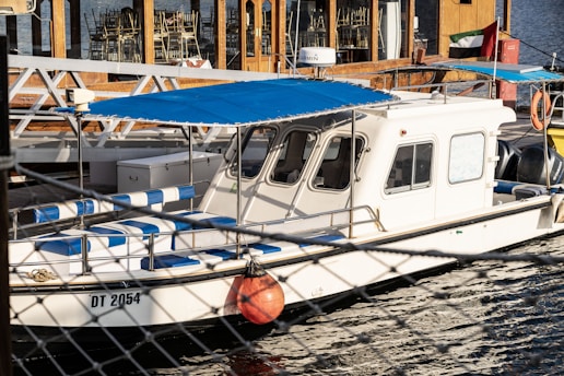 A white boat with a blue canopy is docked by a waterfront. The boat has several blue and white striped seats and a small cabin with windows. In the background, there is a building with wooden elements and stacked chairs. An orange buoy is visible on the side of the boat.