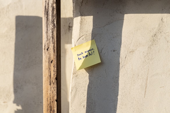 A yellow sticky note with handwritten text saying 'Don't expect, be hopeful!' is attached to a textured, beige wall. The wall is partially in shadow, with the shadow of a wooden post visible on the left side.