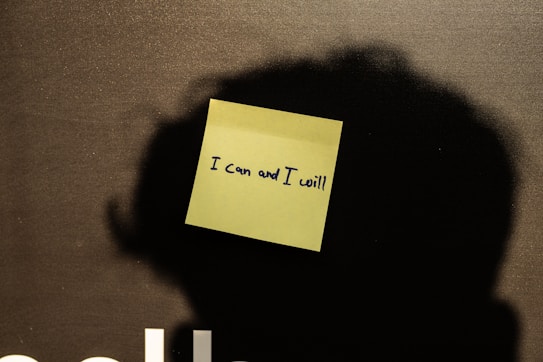 A yellow sticky note with the handwritten motivational phrase 'I can and I will' is placed on a dark surface. The lighting creates a soft shadow around the note.