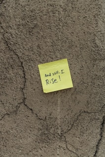 A yellow sticky note with the handwritten message 'And still, I Rise!' is attached to a rough, textured, cracked wall.