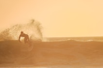 A surfer skillfully rides a wave under the warm glow of a setting sun, creating a dramatic silhouette against the golden sky. The water forms a beautiful spray as the surfer carves through the wave, capturing the dynamic essence of the ocean.