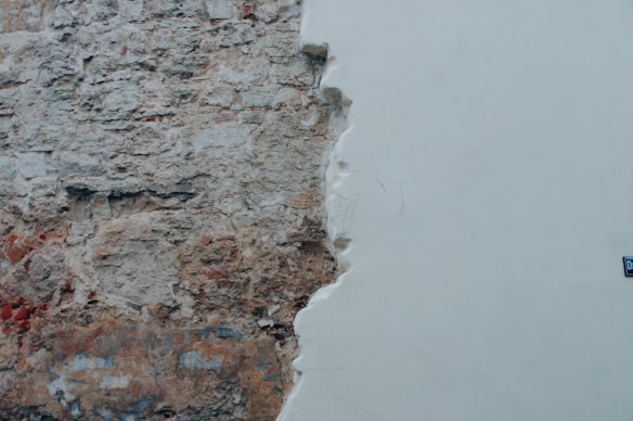 A wall with one half covered in white plaster and the other half showing exposed, weathered bricks and stone. The plastered section appears smooth and clean, while the exposed section reveals the rough texture of the underlying materials. There is a small blue street sign attached to the plastered area.