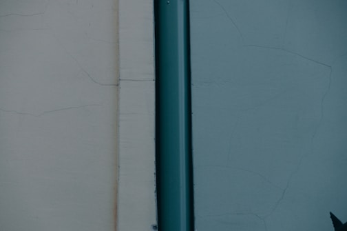 A simple, minimalistic composition featuring a vertical metal pipe on a textured wall. The wall has two sections, with contrasting colors on each side of the pipe, one light beige and the other muted teal. Slight cracks are visible across the surface of the wall, adding an element of aged wear to the scene.