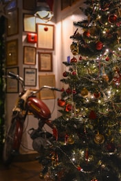 A motorcycle is parked indoors next to a decorated Christmas tree adorned with colorful ornaments and twinkling lights. The background includes framed certificates or documents on the wall, suggesting a cozy indoor setting.