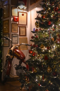 A motorcycle is parked indoors next to a decorated Christmas tree adorned with colorful ornaments and twinkling lights. The background includes framed certificates or documents on the wall, suggesting a cozy indoor setting.