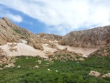 Rocky mountains with patches of snow are surrounded by green vegetation under a partially cloudy sky.
