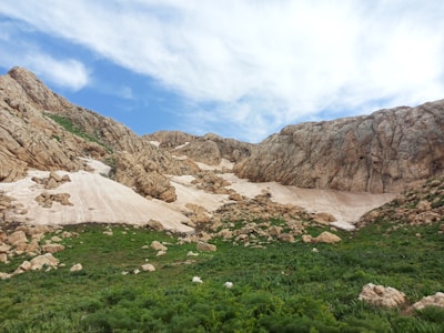 Rocky mountains with patches of snow are surrounded by green vegetation under a partially cloudy sky.