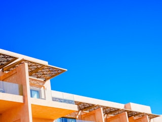 A modern architectural structure with an angular, geometric design set against a vivid blue sky. The building features light, peach-colored walls and large, glass windows, with a decorative lattice roof overhanging some of the windows.