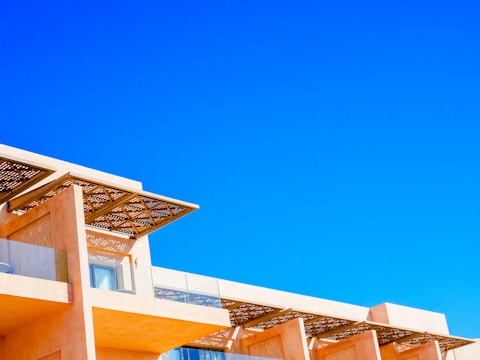 A modern architectural structure with an angular, geometric design set against a vivid blue sky. The building features light, peach-colored walls and large, glass windows, with a decorative lattice roof overhanging some of the windows.