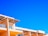 A modern architectural structure with an angular, geometric design set against a vivid blue sky. The building features light, peach-colored walls and large, glass windows, with a decorative lattice roof overhanging some of the windows.