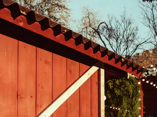 A rustic, red wooden structure with a slanted roof is adorned with greenery and festive string lights. The sky is clear, and bare trees are visible in the background, suggesting a seasonal setting.