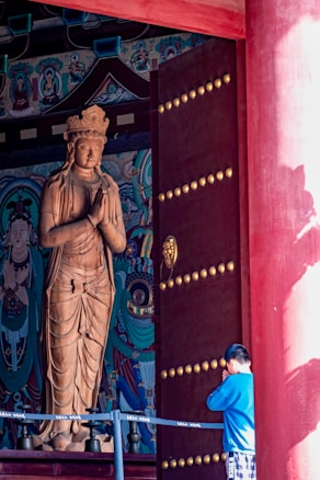 A large wooden statue of a deity stands inside a decorated temple, surrounded by colorful intricate artwork on the walls. A young person in a blue top and plaid shorts stands with hands clasped together in a prayerful gesture facing the statue. The atmosphere is serene, with red columns and a partially open door with golden embellishments.