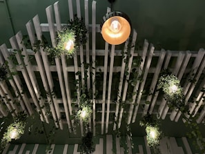 A ceiling with white slatted panels features hanging green foliage and several warm light bulbs integrated into the design, creating a modern, natural aesthetic.