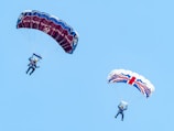 Two parachutists are descending against a clear blue sky. The parachute on the left is dark blue with red accents, while the one on the right features a white background with a red and blue Union Jack pattern. Both parachutists are wearing blue jumpsuits and helmets.