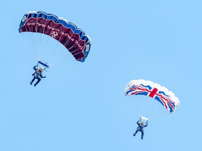 Two parachutists are descending against a clear blue sky. The parachute on the left is dark blue with red accents, while the one on the right features a white background with a red and blue Union Jack pattern. Both parachutists are wearing blue jumpsuits and helmets.