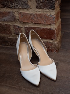 A pair of elegant white high-heeled shoes is placed on a wooden floor next to a rustic brick wall. The shoes have a pointed toe design and are decorated with subtle metallic elements along the edges.
