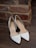 A pair of elegant white high-heeled shoes is placed on a wooden floor next to a rustic brick wall. The shoes have a pointed toe design and are decorated with subtle metallic elements along the edges.
