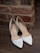 A pair of elegant white high-heeled shoes is placed on a wooden floor next to a rustic brick wall. The shoes have a pointed toe design and are decorated with subtle metallic elements along the edges.