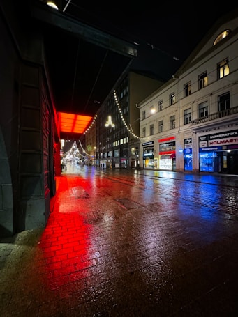 A dimly lit city street at night with wet pavement reflecting red and blue lights. Buildings line the right side, and strings of white lights hang above the street. A prominent red light glows from an awning on the left.