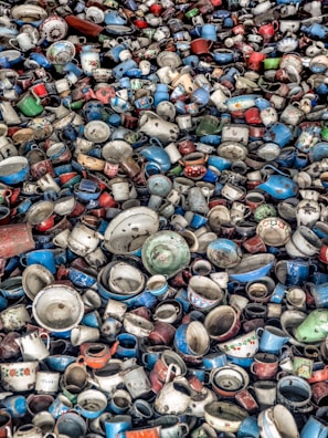 A large collection of vintage and colorful enamel mugs, pots, and kitchenware tightly packed together. The dishware is a mix of various shades including blue, red, white, and green, with some showing floral patterns or labels.