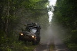 A rugged off-road vehicle is navigating a narrow, muddy path surrounded by dense green forest. The vehicle is loaded with gear on its roof, indicating an adventurous journey. Mist or dust creates a hazy atmosphere, adding to the sense of wilderness.