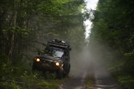 A rugged off-road vehicle is navigating a narrow, muddy path surrounded by dense green forest. The vehicle is loaded with gear on its roof, indicating an adventurous journey. Mist or dust creates a hazy atmosphere, adding to the sense of wilderness.
