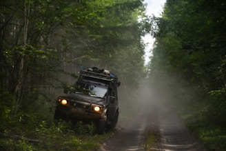 A rugged off-road vehicle is navigating a narrow, muddy path surrounded by dense green forest. The vehicle is loaded with gear on its roof, indicating an adventurous journey. Mist or dust creates a hazy atmosphere, adding to the sense of wilderness.