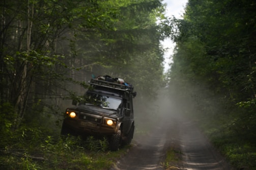 A rugged off-road vehicle is navigating a narrow, muddy path surrounded by dense green forest. The vehicle is loaded with gear on its roof, indicating an adventurous journey. Mist or dust creates a hazy atmosphere, adding to the sense of wilderness.